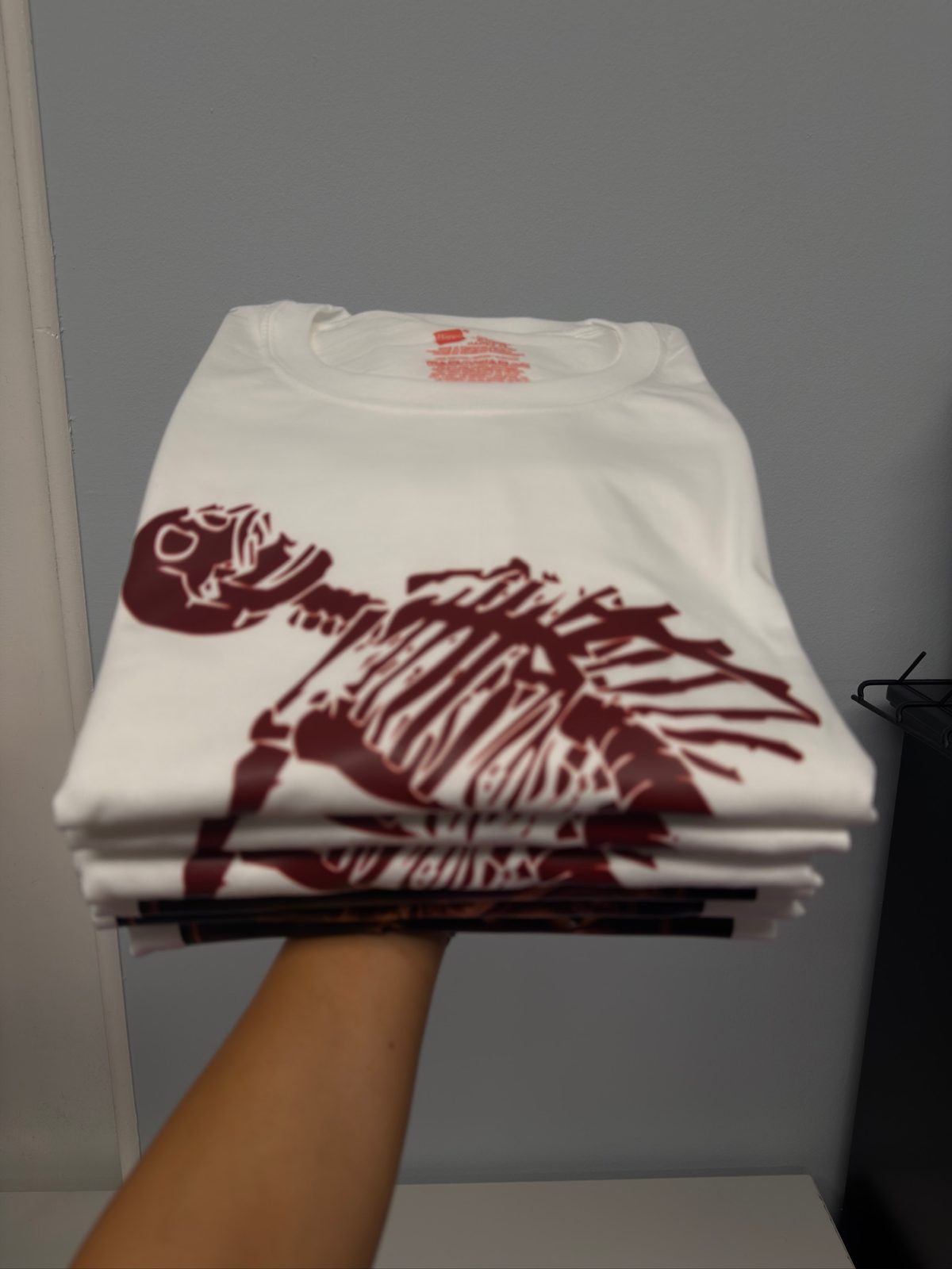 A bold flat lay of freshly printed DTF transfers and vibrant custom t-shirts spread across a clean, dark workbench. The transfers feature colorful graphics, gradients, and intricate details on glossy transparent film, their edges curling slightly. Beside them, a heat press stands open, its platen gleaming, with a neatly aligned blank hoodie ready for pressing. Soft studio lighting from above creates crisp highlights on the film and subtle shadows on the apparel, emphasizing texture in the cotton fabric. The background is slightly blurred shelves stacked with rolls of film, ink bottles, and neatly labeled boxes, evoking an efficient print garage. Photographic realism, eye-level angle, sharp focus on the transfers, with a bold, energetic mood that highlights professional, small-shop craftsmanship.